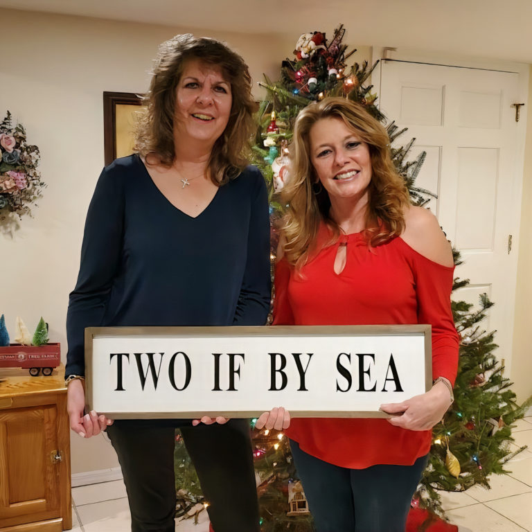 Maureen and Danielle holding up a sign that says "Two If By Sea" in front of a christmas tree